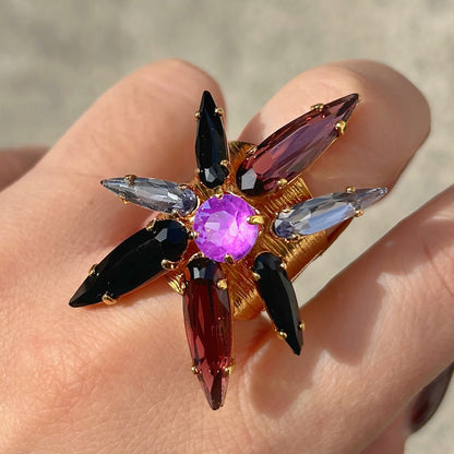 Bague Astra Mûre Crézus en forme d'étoile avec cristal central rond violet orchidée, huit cristaux pointus noirs, prune et bleu lavande sur large monture dorée texturée.