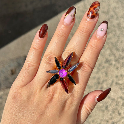 Bague Astra Mûre Crézus en forme d'étoile avec cristal central rond violet orchidée, huit cristaux pointus noirs, prune et bleu lavande sur large monture dorée texturée.