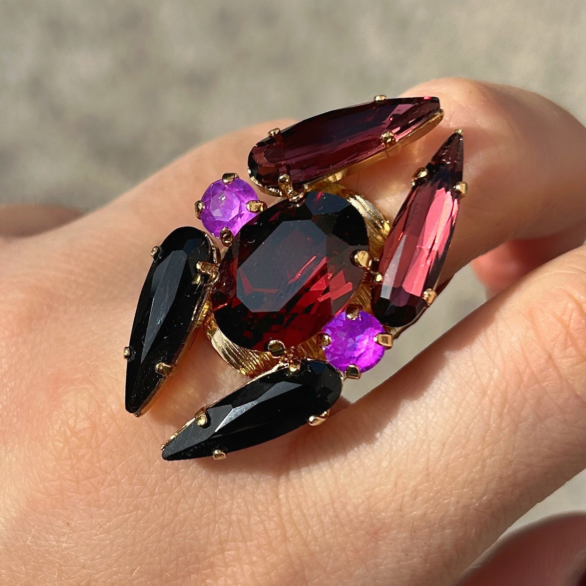 Bague réglable Lumen Mûre Crézus avec grand cristal ovale grenat, cristaux noirs et prune allongés, deux cristaux ronds violet orchidée et monture dorée texturée.