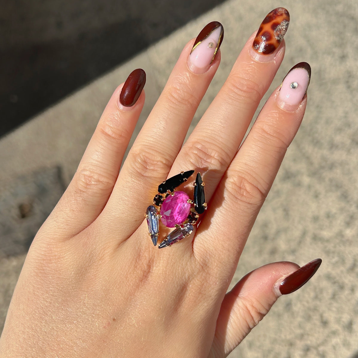 Bague Mira Mûre Crézus avec grand cristal ovale rose fuchsia, cristaux pointus noirs et bleu lavande, petits cristaux prune et monture dorée à anneau large.