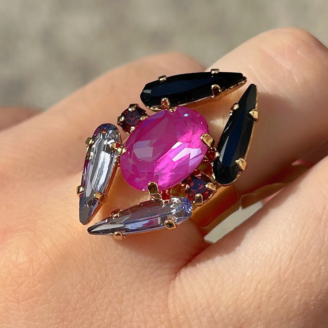Bague Mira Mûre Crézus avec grand cristal ovale rose fuchsia, cristaux pointus noirs et bleu lavande, petits cristaux prune et monture dorée à anneau large.