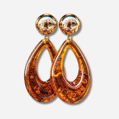 Boucles d’oreilles à clips Céleste Ambre Francine Bramli Paris, avec cabochon rond doré effet miroir et grand pendentif goutte ajourée en résine ambre translucide aux inclusions foncées.
