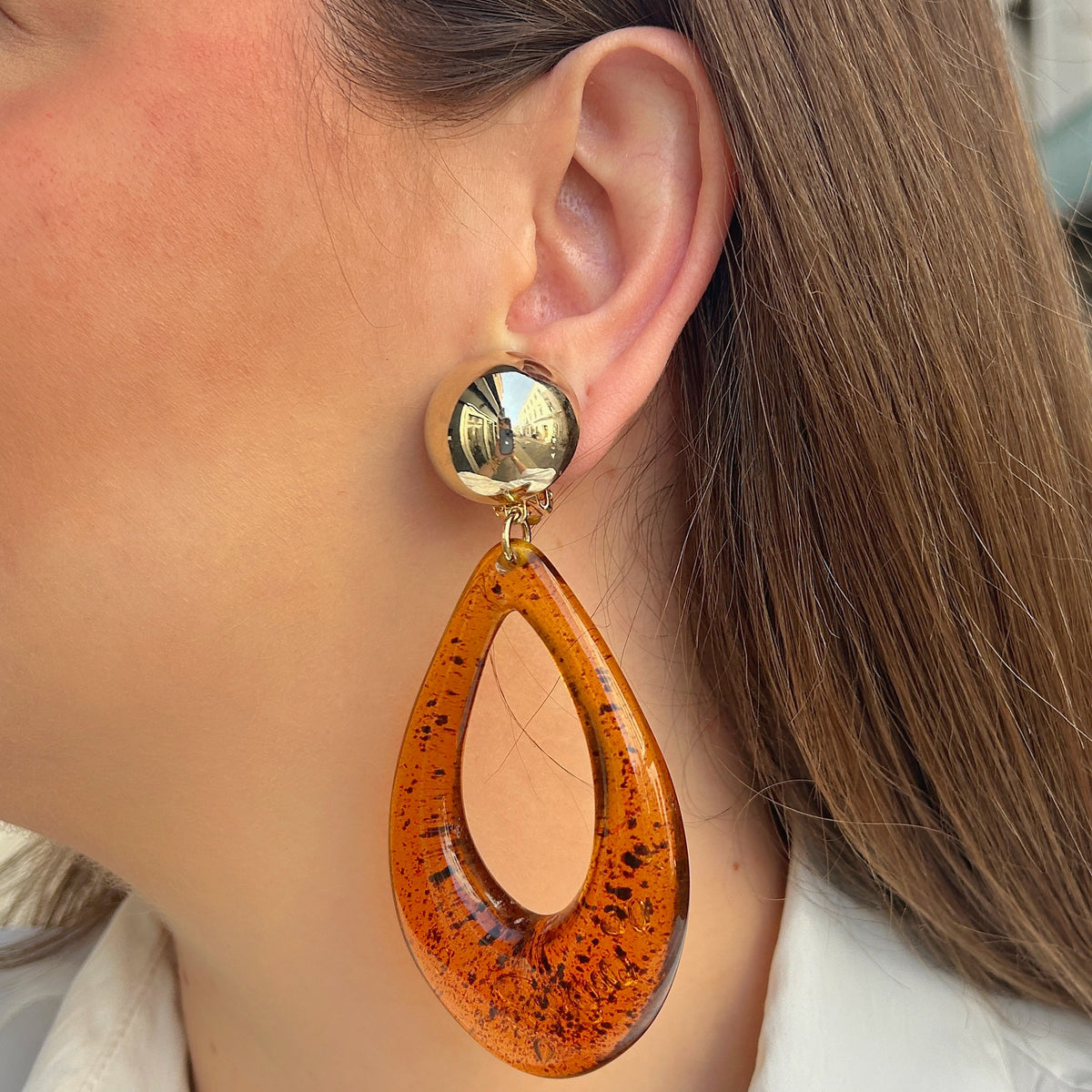 Boucles d’oreilles à clips Céleste Ambre Francine Bramli Paris, avec cabochon rond doré effet miroir et grand pendentif goutte ajourée en résine ambre translucide aux inclusions foncées.