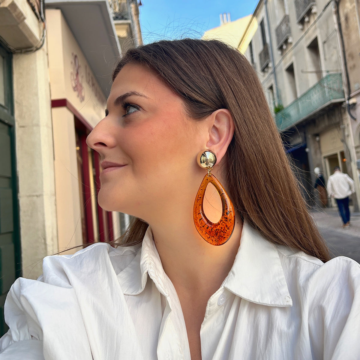 Boucles d’oreilles à clips Céleste Ambre Francine Bramli Paris, avec cabochon rond doré effet miroir et grand pendentif goutte ajourée en résine ambre translucide aux inclusions foncées.