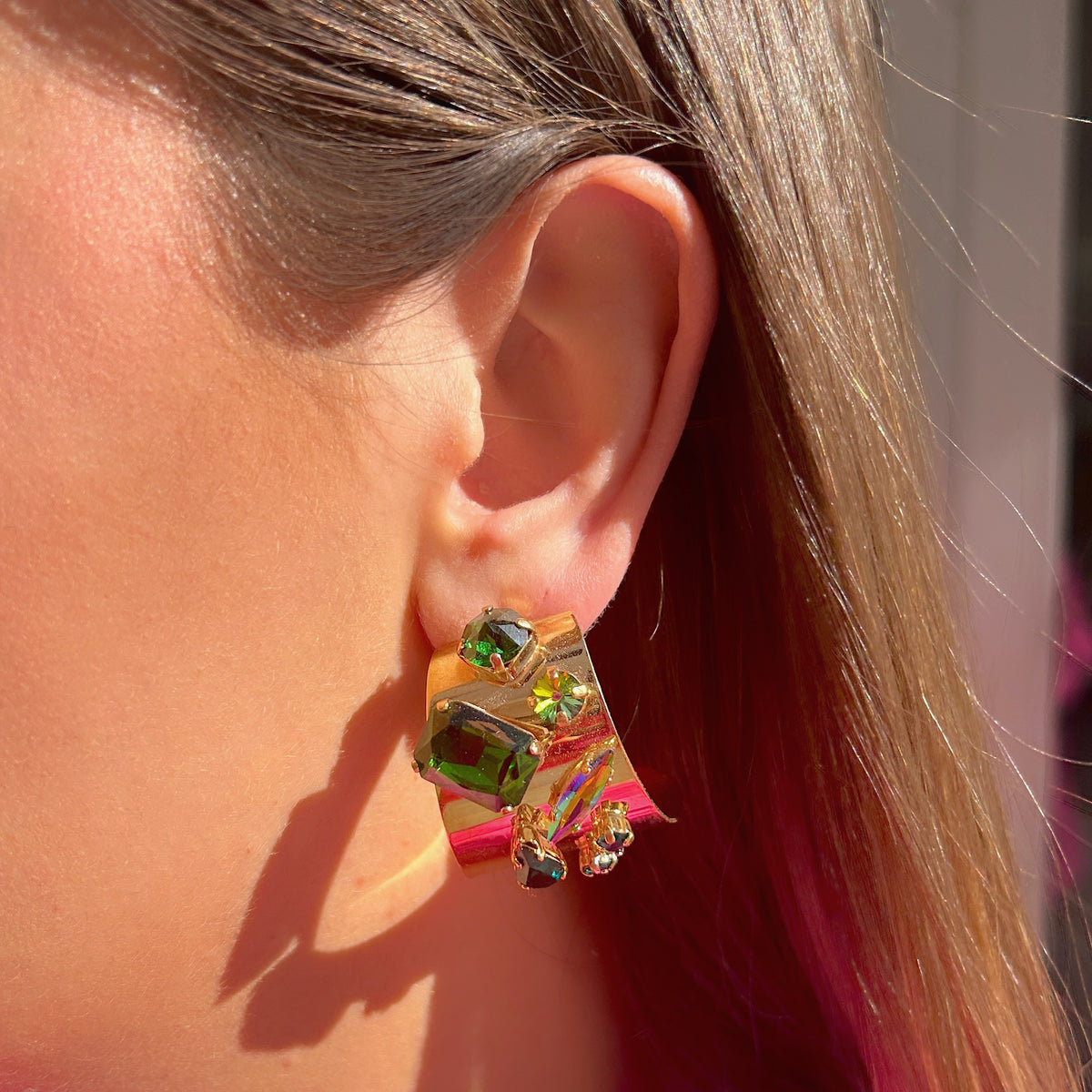Boucles d’oreilles Lumeia Crézus à tige, monture dorée large et incurvée ornée de cristaux facettés vert émeraude, de pierres en goutte et de cristaux multicolores aux reflets roses, violets et bleus.