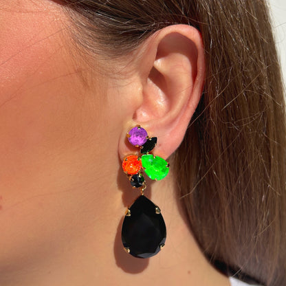 Boucle d’oreille Prisma Fluo Crézus à clips portée à l’oreille, cristaux facettés violet, vert fluo, orange et noir, grande pampille goutte noire sur monture dorée.