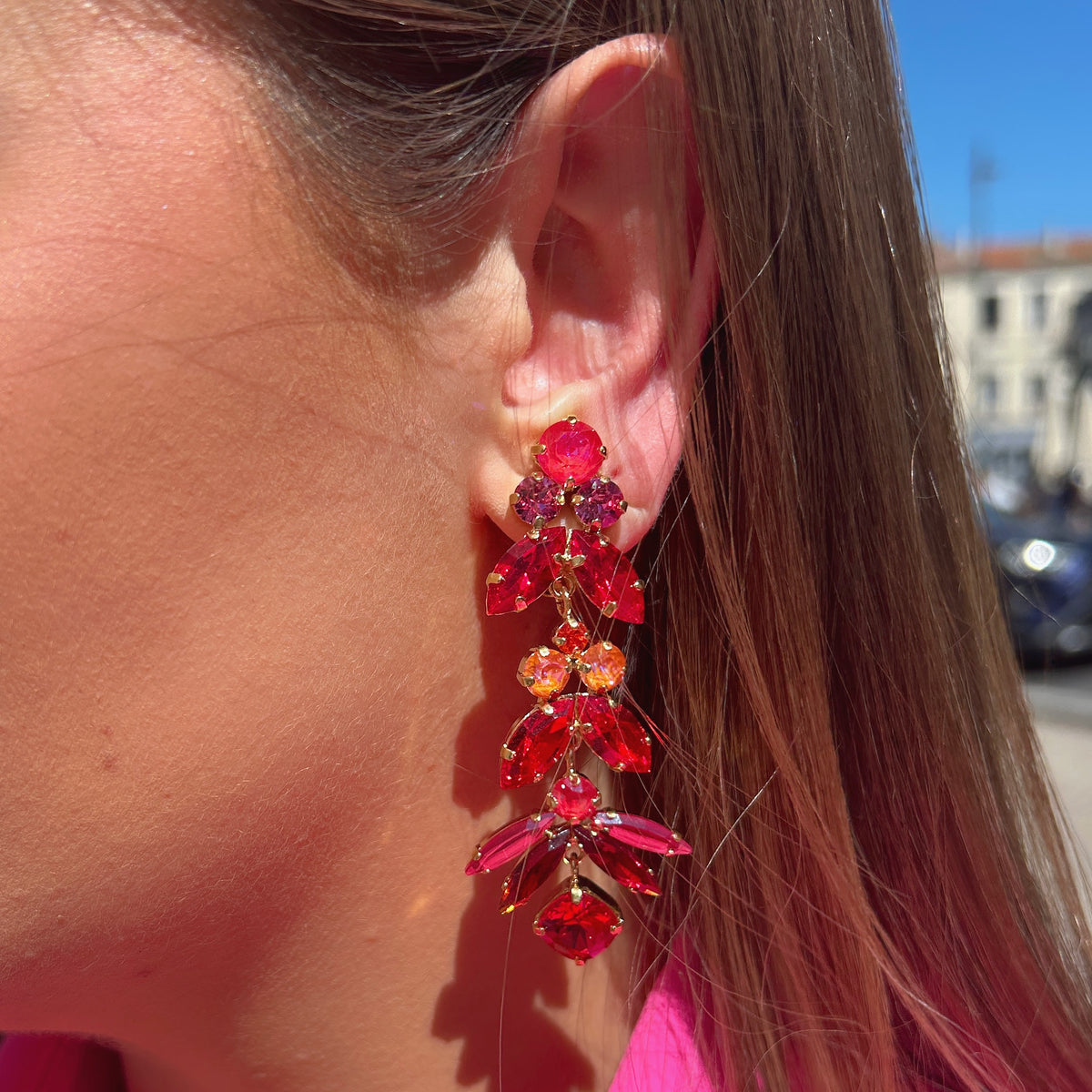 Boucles d’oreilles pendantes Sophéa Red Crézus, monture dorée ornée de cristaux facettés rouge rubis, rose fuchsia et orange, disposés en cascade avec pierres rondes et marquises.