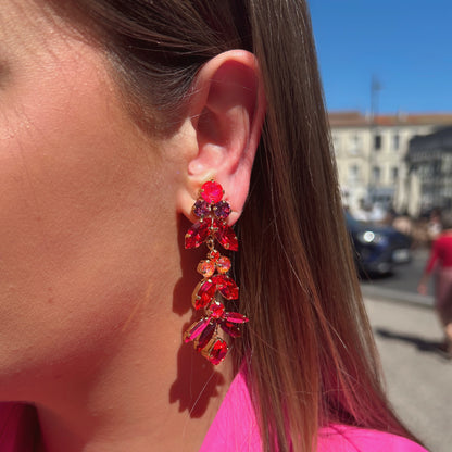 Boucles d’oreilles pendantes Sophéa Red Crézus, monture dorée ornée de cristaux facettés rouge rubis, rose fuchsia et orange, disposés en cascade avec pierres rondes et marquises.